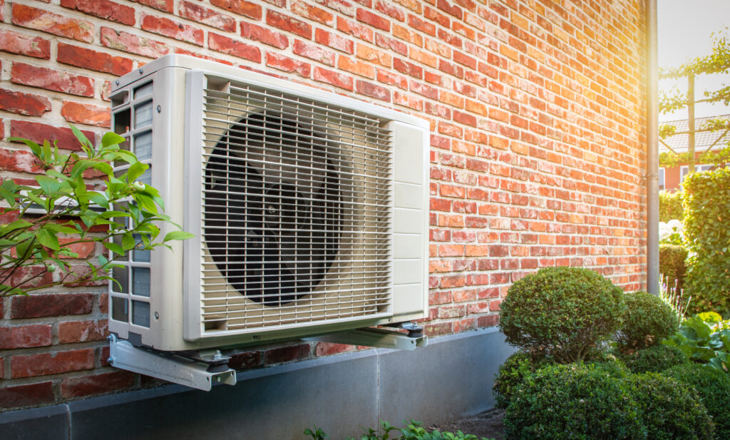 Side view of outdoor energy unit hanging on brick wall of house on a sunny day. Air to air heat pump for cooling or heating the home. Outdoor unit powered by renewable energy. Air conditioner and air heat pump. HVAC Service Near Me